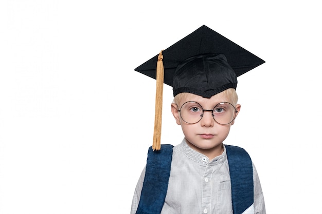 Portret ślicznego blond chłopca w dużych okularach, akademickim kapeluszu i szkolnej torbie.