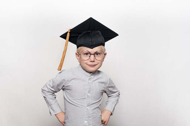 Portret ślicznego blond chłopca w dużych okularach i akademickim kapeluszu. Białe tło.