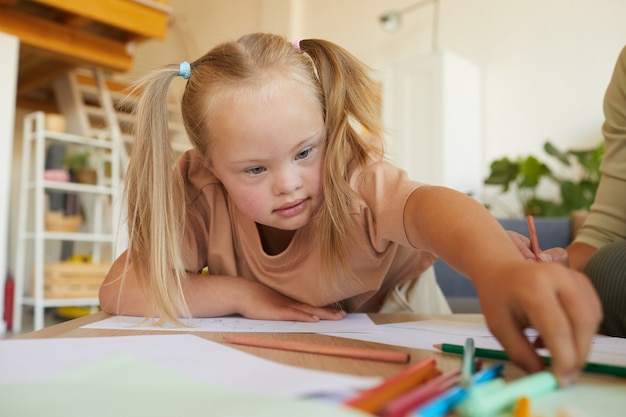 Portret ślicznej blondynki z zespołem Downa, rysująca i sięgająca po ołówki, ciesząc się lekcją rozwoju, kopiuj przestrzeń