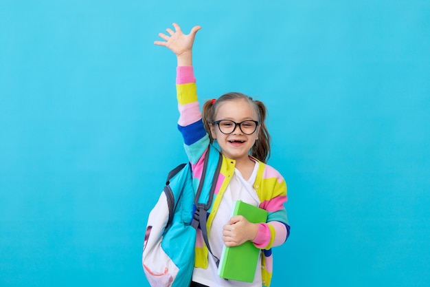 Portret ślicznej dziewczynki w okularach w pasiastej kurtce z notebookami i plecakiem, radując się i bawiąc Koncepcja edukacji Studio fotograficzne niebieskie tło miejsce na tekst