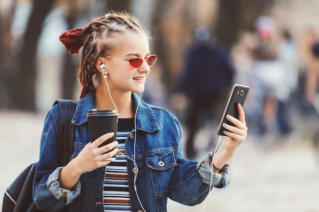 Portret ślicznej dziewczyny hipster z dredami w słuchawkach i okularach, pije kawę i śmieje się, przesuwając strony na swoim smartfonie