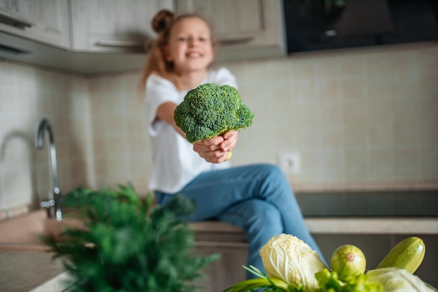 Portret ślicznej małej dziewczynki w kuchni z zieloną kapustą i brokułami, świeżymi warzywami i sałatką