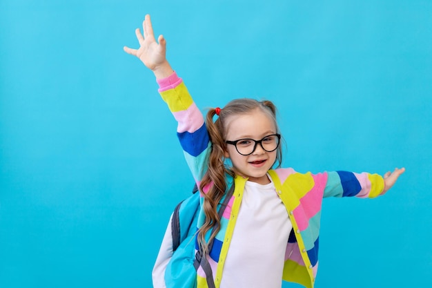 Portret ślicznej małej dziewczynki w okularach w pasiastej kurtce z zeszytami i plecakiem, ciesząc się i bawiąc. Pojęcie edukacji. Studio fotograficzne, niebieskie tło, miejsce na tekst.