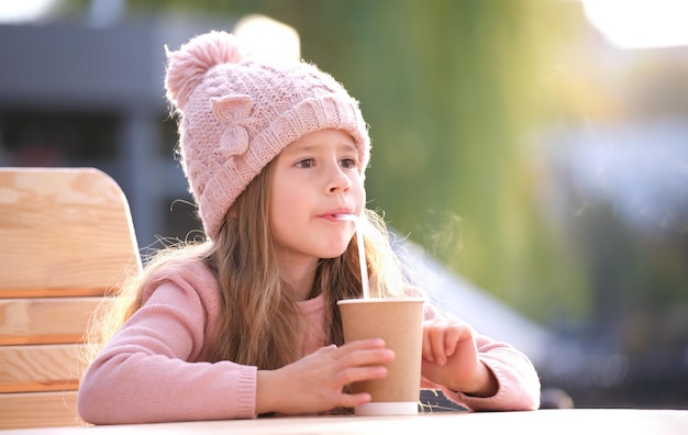Portret ślicznej małej dziewczynki w różowym kapeluszu, siedzącej samotnie w ulicznej kawiarni, pijącej herbatę z papierowego kubka