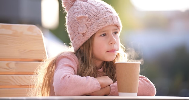 Portret ślicznej małej dziewczynki w różowym kapeluszu, siedzącej samotnie w ulicznej kawiarni, pijącej herbatę z papierowego kubka