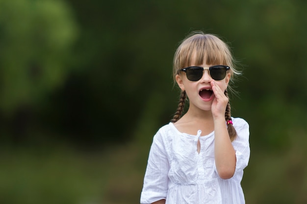 Portret ślicznej ślicznej śmiesznej bezzębnej modnej młodej dziewczyny z blond warkoczami w białej sukni i ciemnych okularach przeciwsłonecznych, nazywających kogoś