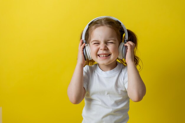 Portret ślicznej wesołej małej dziewczynki słuchania muzyki z białymi słuchawkami. audiobooki, lekcje audio. koncepcja edukacji. studio fotograficzne, żółte tło, miejsce na tekst. Zdjęcie wysokiej jakości