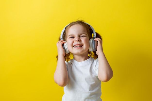 Portret ślicznej wesołej małej dziewczynki słuchania muzyki z białymi słuchawkami. audiobooki, lekcje audio. koncepcja edukacji. studio fotograficzne, żółte tło, miejsce na tekst. Zdjęcie wysokiej jakości