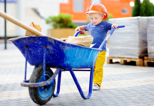 Zdjęcie portret śliczny berbecia budowniczy w hardhats z taczką pracuje outdoors.