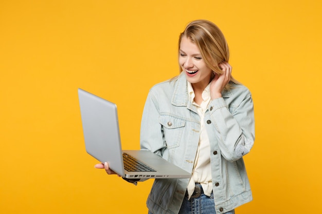 Portret śmiejącej się radosnej młodej kobiety w dżinsowych ubraniach casualowych, przy użyciu komputera typu laptop na białym tle na żółto pomarańczowej ścianie w studio. Koncepcja życia ludzi. Makieta miejsca na kopię.