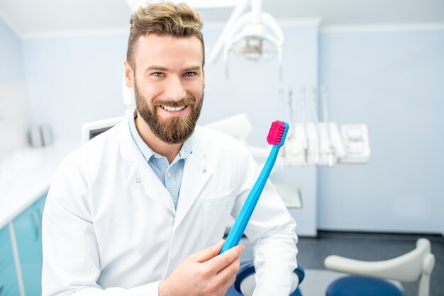 Portret śmiesznego dentysty w mundurze z dużą szczoteczką do zębów w gabinecie stomatologicznym