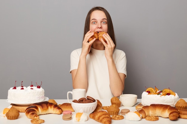 Portret śmiesznej głodnej kobiety z brązowymi włosami w białej koszulce siedzącej przy stole pełnym słodkich deserów łamiących dietę gryzie dwa rogaliki w tym samym czasie gryząc odizolowane na szarym tle