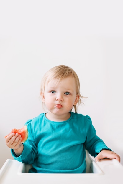 Portret śmiesznej małej dziewczynki siedzącej na wysokim krześle i jedzącej arbuza