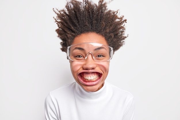 Portret śmiesznej pozytywnej kobiety o ciemnej karnacji uśmiecha się z zębami ma duże pozy pod ciśnieniem powietrza ma włosy stojące ubrane w swobodny poloneck i duże przezroczyste okulary