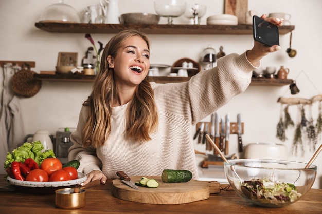 portret śmiesznej słodkiej kobiety w zwykłych ubraniach, biorącej selfie na telefon i śmiejącej się w przytulnej kuchni