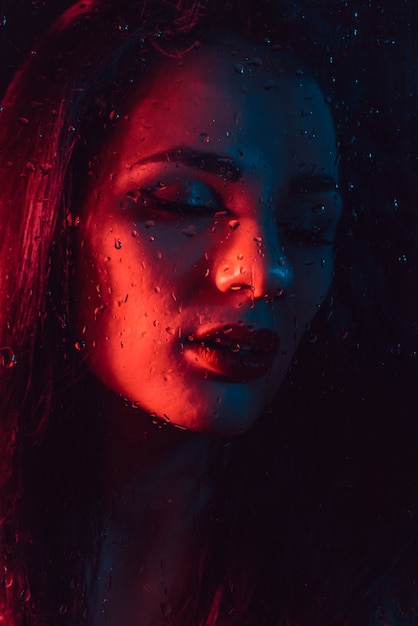 Zdjęcie portret smutna młoda dziewczyna z czerwonym błękitnym oświetleniem za szkłem z raindrops