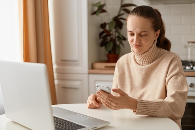 Portret smutnej rozczarowanej młodej kobiety w beżowym swetrze, siedzącej w kuchni z notatnikiem pracującym online i używającej telefonu, czytającego złe wieści