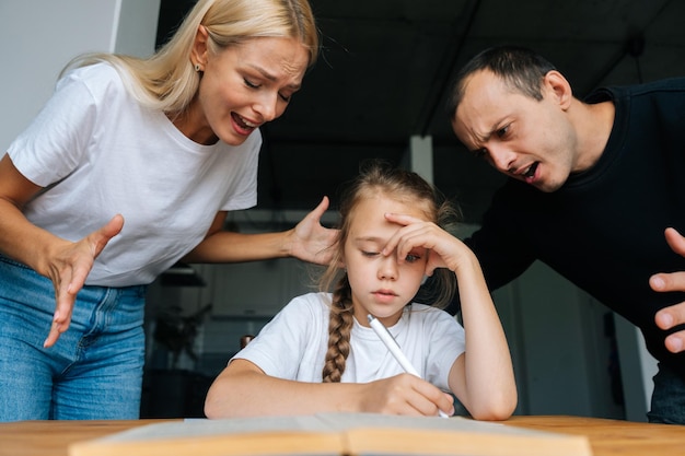 Portret smutnej, słodkiej dziewczynki piszącej pracę domową siedzącej przy stole na tle wściekłych rodziców krzyczących i karcących razem leniwą córkę