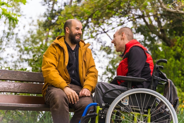 Portret sparaliżowanego młodego mężczyzny na wózku inwalidzkim z przyjacielem na ławce w publicznym parku w mieście, rozmawiając i śmiejąc się