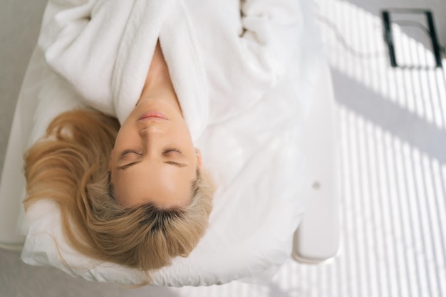Portret spokojnej młodej kobiety w białym szlafroku leżącej z zamkniętymi oczami na kanapie medycznej po zabiegu spa Zbliżenie blondynki relaksującej się w nowoczesnej klinice piękności