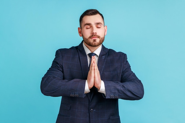 Portret spokojny, zrelaksowany młody człowiek z brodą, ubrany w garnitur w oficjalnym stylu, stojący z podniesionymi rękami i wykonujący ćwiczenia jogi medytacji Kryty studio strzał na białym tle na niebieskim tle