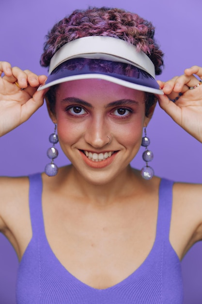 Portret sportowej mody kobiety uśmiechającej się zębami do kamery ze stylowymi kolczykami w uszach w fioletowym dresie do jogi i przezroczystą czapką na fioletowym monochromatycznym tle