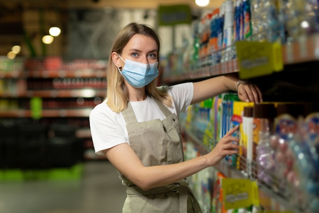 Portret sprzedawczyni, w masce ochronnej kobieta uśmiecha się i patrząc na kamery w supermarkecie. Przyjemna przyjazna sprzedawczyni stojąca w sklepie między rzędami. pracownik ze skrzyżowanymi rękami