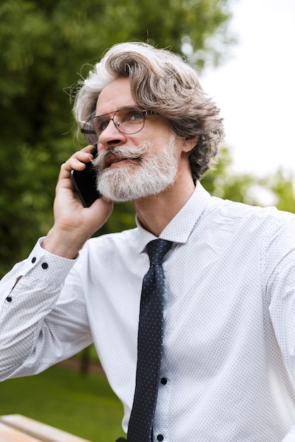 Portret Starego Biznesmena W Okularach Rozmawiającego Przez Telefon, Siedząc Na ławce W Letnim Parku