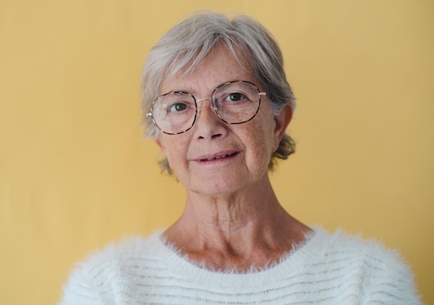 Portret starej atrakcyjnej starszej kobiety w okularach i białym swetrze patrzącej na kamerę, na żółtym tle