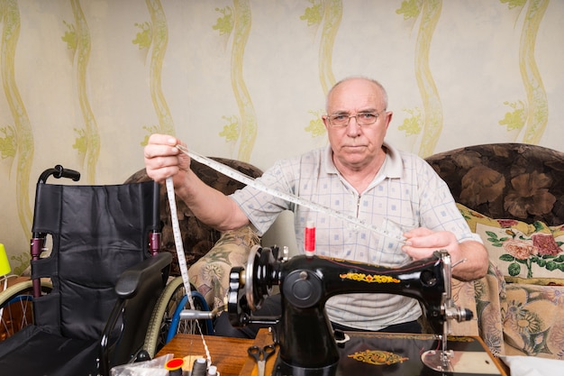 Portret starszego mężczyzny trzymającego taśmę mierniczą i siedzącego w staromodnej ręcznej maszynie do szycia obok wózka inwalidzkiego w salonie w domu i patrząc na kamerę