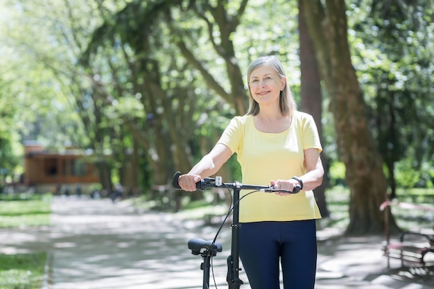 Portret starszej aktywnej kobiety siwowłosa kobieta w parku patrząca na kamerę i uśmiechająca się z rowerowym letnim dniem