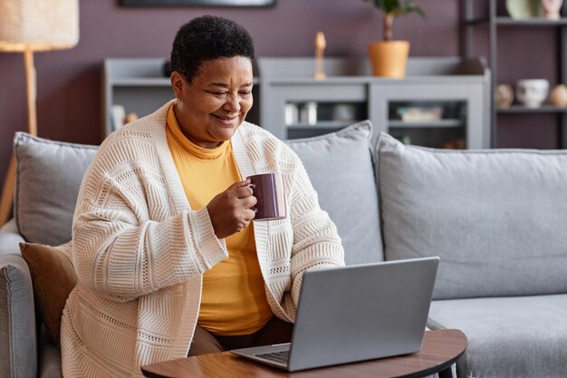 Portret starszej czarnej kobiety używającej laptopa w domu i cieszącej się filiżanką kawy
