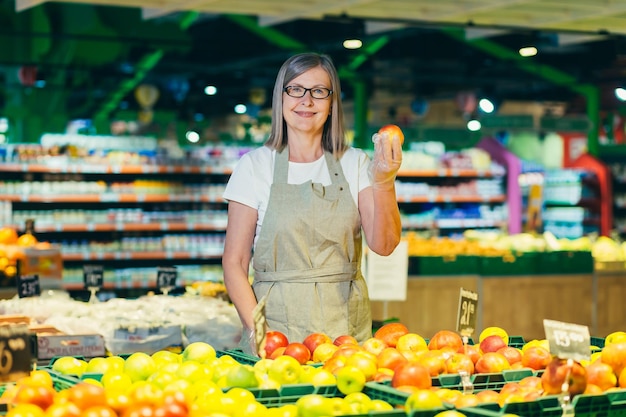 Portret starszej kobiety w okularach w supermarkecie pracowniczym sklepu spożywczego Stoi w ubraniach roboczych w pobliżu owoców i ofochi pokazuje jedzenie patrzy w kamerę się uśmiecha