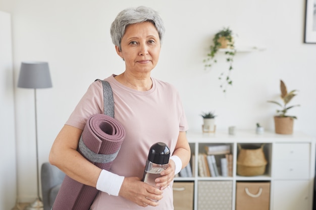 Portret starszej kobiety z siwymi włosami, trzymając butelkę wody i matę do ćwiczeń