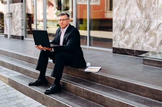 Portret Starszy Mężczyzna Siedzi Otwartego Laptop Na Zewnątrz I Trzyma W Kostiumu.