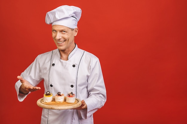 Portret starszy piekarz trzyma pyszne ciasta na białym tle na czerwonym tle.