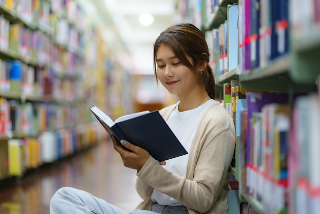 Portret studenta Uniwersytetu Smart Asian kobieta czytanie książki i patrząc na kamery między półkami w bibliotece kampusu z copyspace.