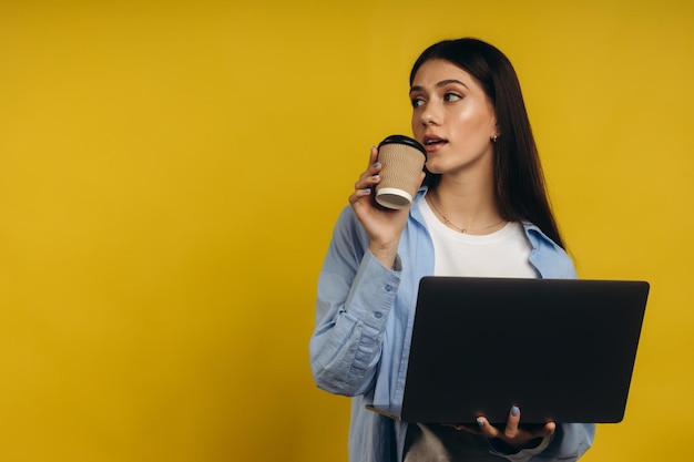 Portret studentki trzymającej filiżankę kawy podczas pracy na laptopie na odosobnionym żółtym tle