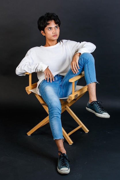 Portret studio całego ciała strzał azjatyckich młodych LGBTQ gejów glamour przystojny biseksualnych homoseksualnych mężczyzn model w swobodnym stroju siedzi skrzyżowane nogi na drewnianym krześle pozowanie, gestykulując na czarnym tle