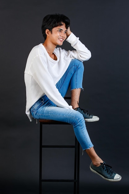 Portret studio całego ciała strzał azjatyckich młodych LGBTQ gejów glamour przystojny mężczyzna modelu w casual biała koszula dżinsy trampki siedząc na wysokim krześle, śmiejąc się, trzymając rękę okładka usta na czarnym tle