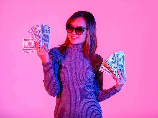 Portret studio strzał azjatyckich młodych bogatych bogatych fajne modelki w szary sweter z golfem i okulary przeciwsłoneczne z czarnymi obiektywami, trzymając pełny stos waluty banknotów sto dolarów na różowym tle światła.
