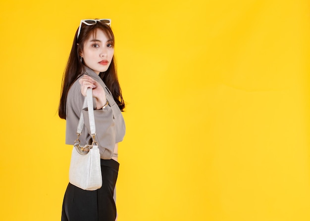 Portret studio strzał azjatyckich modne modne kobiece hipster nastolatek model w casual crop top ulicy nosi kurtkę okulary przeciwsłoneczne trampki trzymając torebkę torebkę spójrz na aparat na żółtym tle