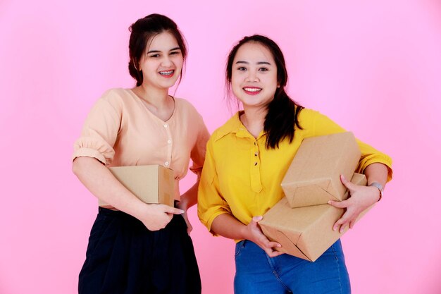 Portret studio strzał dwóch azjatyckich pulchne i smukłe szelki zęby kucyk włosy partnerka biznesowa w casual stojącej uśmiechnięta trzymająca dostawę paczki razem na różowym tle.