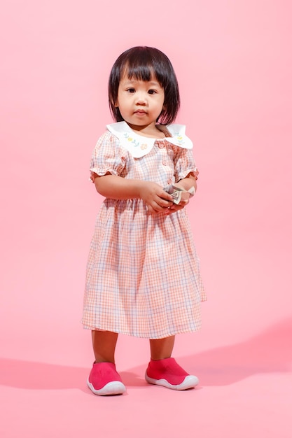 Portret studio strzał mały ładny azjatycki przedszkole przedszkole dziewczyna córka model w dorywczo długa sukienka i buty stojące patrzeć na aparat trzymając banknot stu dolarów w ręce na różowym tle.