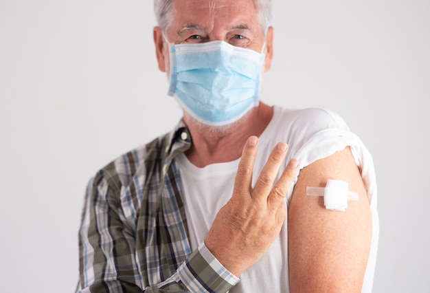 Portret studio strzał starego kaukaskiego starszego mężczyzny pacjenta noszącego maskę pokazującą palcami numer trzy jako trzecie dawki szczepionki na koronawirusa Covid-19, patrząc na kamerę
