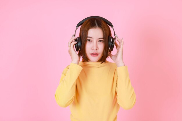Portret studio wyłącznik strzał azjatyckich młodych dość krótkie włosy modelki w żółtą koszulę z długim rękawem, stojąc trzymając dostosowując duże czarne słuchawki podczas słuchania muzyki na różowym tle.
