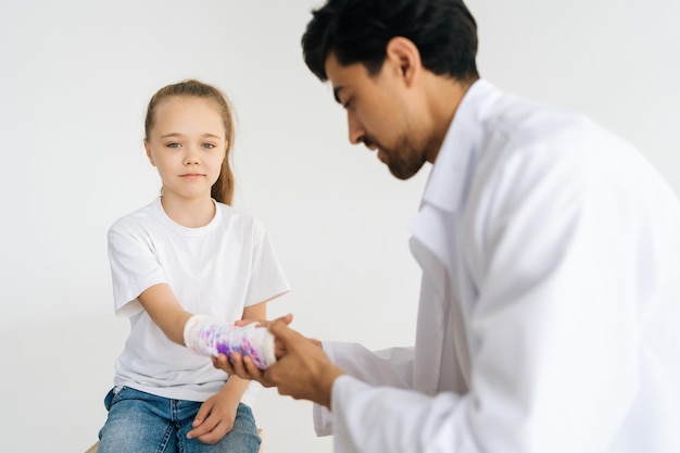 Portret studyjny ślicznej blondynki małej dziewczynki ze złamaną ręką owiniętą w biały bandaż gipsowy otrzymującej opiekę medyczną lekarza pediatry