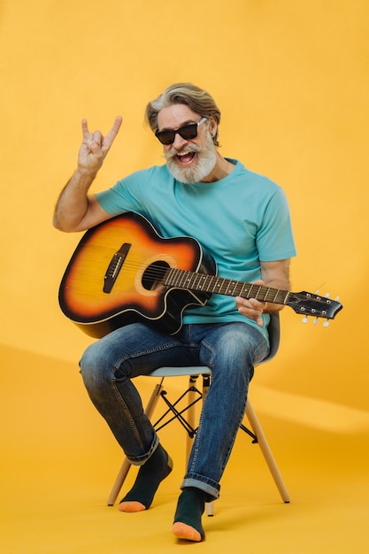 Portret studyjny starszego siwego mężczyzny w okularach przeciwsłonecznych grającego na gitarze