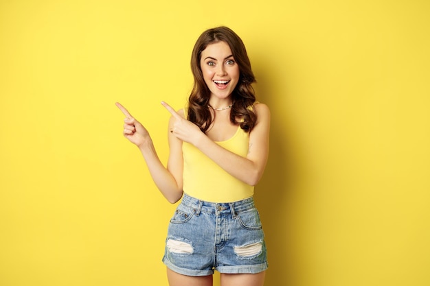 Portret stylowe lato dziewczyna, brunetka kobieta pokazując drogę, wskazując palcami w lewo, reklama, stojąc na żółtym tle. Skopiuj miejsce