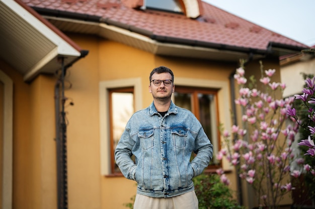 Zdjęcie portret stylowego mężczyzny w okularach i dżinsowej kurtce postawiony na tle jego domu